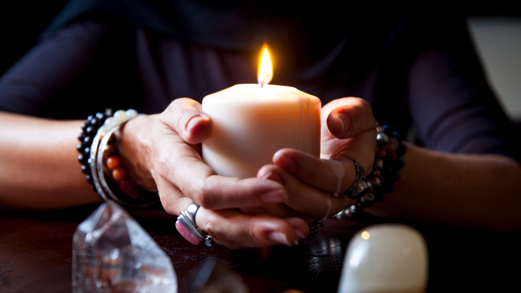 A single lit candle in a dimly lit room, casting a warm glow. The candle is surrounded by autumn leaves and small stones, creating a cozy, reflective atmosphere reminiscent of the Samhain season. Soft shadows enhance the feeling of calm and introspection.